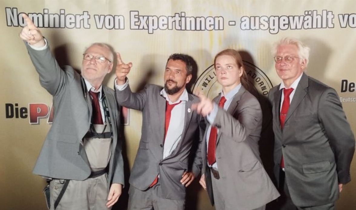 Vier Personen in grauen Anzügen mit roten Krawatten vor einer Fotowand, drei von ihnen zeigen in unterschiedliche Richtungen. Auf der Wand das Logo von Die PARTEI, das Siegel der T. Willibald Adorno Stiftung und der Schriftzug „Nominiert von Expertinnen – ausgewählt vo…“.